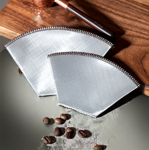 Två trapetsformade kaffefilter i finmaskigt rostfritt stål med sydd kant, liggande platt bredvid kaffebönor